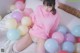 A woman in a pink sweatshirt sitting on a bed surrounded by balloons.